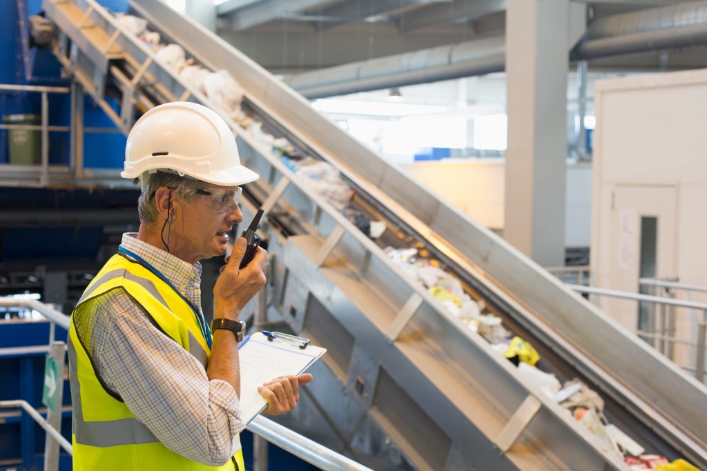 A man in a hard hat and safety vest speaking into a walkie talkie inside recycling facility.